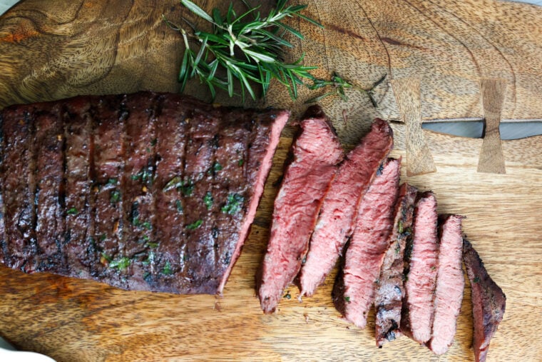 A juicy grilled steak, cooked medium rare with a pink center and crispy char on the outside, sits partly sliced on a wooden cutting board. Theres even a fresh sprig of rosemary next to it for that extra touch.