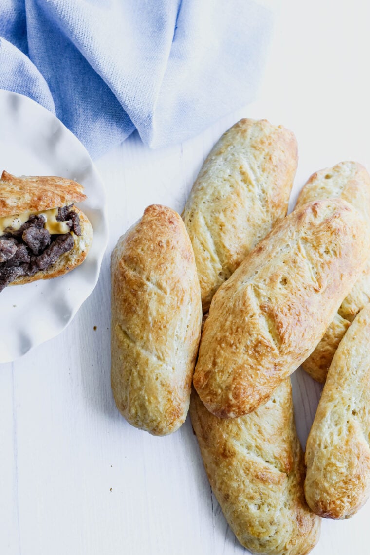 Five golden-brown hoagie buns are lined up on a white surface, right by a plate with an open roll stuffed with beef and melty cheese. There’s also a light blue cloth peeking in the background.