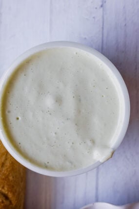 Here’s a close-up shot from above: a white bowl packed with creamy jalapeno aioli sitting on a light wooden table, with a bit of bread peeking in at the edge.