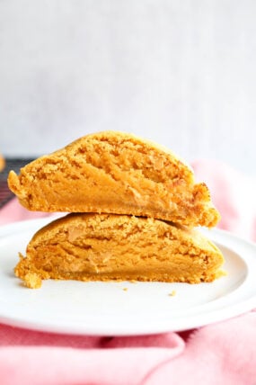 Two big, golden-brown cookies are stacked on a white plate, each one broken in half to show off their soft, dense insides. There’s a pink cloth in the background.