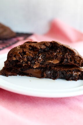 A gooey, chocolate-packed cookie cracked in half on a white plate, showing off that melty chocolate center. In the background, there’s a soft pink cloth and a cooling rack.