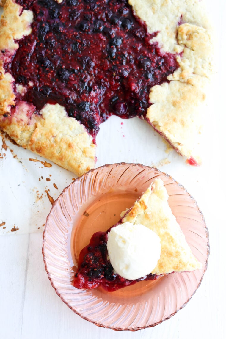 A slice of berry galette with a scoop of vanilla ice cream sits on a pink glass plate, right next to the rest of the galette on a white table.