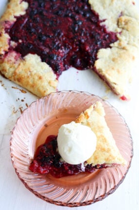 A slice of mixed berry galette with a scoop of vanilla ice cream is on a pink glass plate, while the rest of the cozy-looking galette chills in the background on some parchment paper.