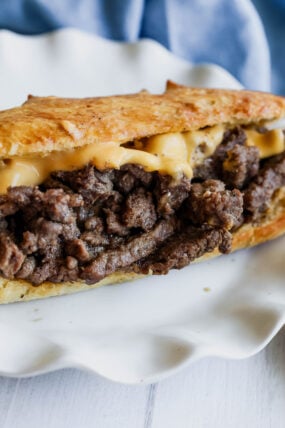 Philly cheesesteak vibes: juicy seasoned beef and gooey melted cheese stuffed in a hoagie roll, all served up on a white plate. Blue cloth chilling in the background!.