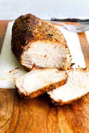 A juicy, roasted pork loin is chilling on a wooden cutting board, partly sliced to show off just how tender it is. Two thick pieces are up front, with a fork and knife hanging out in the background.