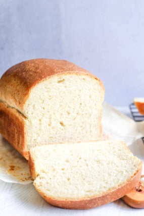 A loaf of white bread rests on parchment paper, with a chunky slice cut and laid out in front. The crust is golden brown, and the inside looks super soft and fluffy. The background’s simple and light.