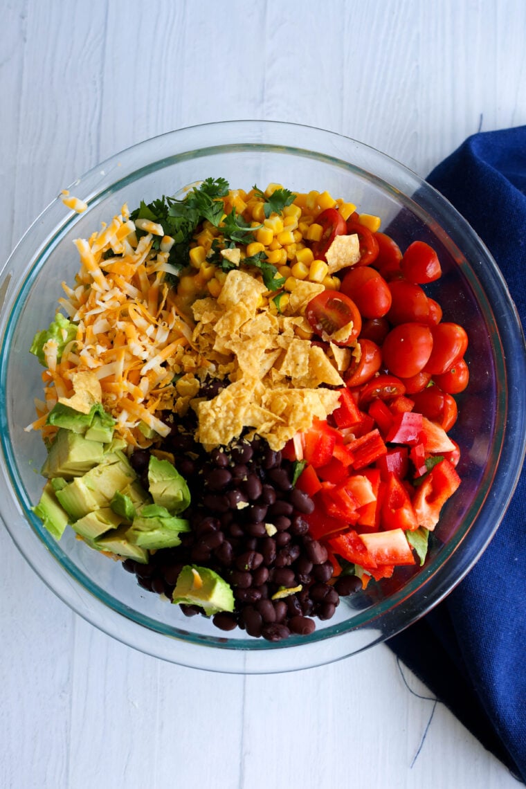A glass bowl packed with black beans, avocado chunks, shredded cheese, red bell pepper pieces, cherry tomatoes, corn, cilantro, and some crushed tortilla chips sits on a white surface next to a navy blue napkin.