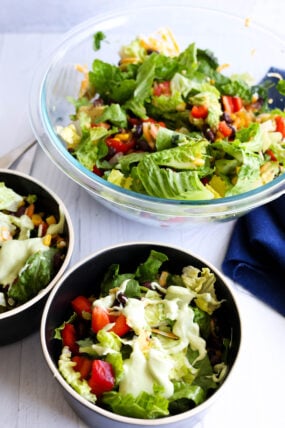 A big glass bowl and two smaller ones are loaded with mixed greens, tomatoes, black beans, corn, and creamy dressing on top. They’re all sitting on a white surface next to a blue napkin.