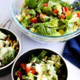 A big glass bowl and two smaller ones are loaded with mixed greens, tomatoes, black beans, corn, and creamy dressing on top. They’re all sitting on a white surface next to a blue napkin.