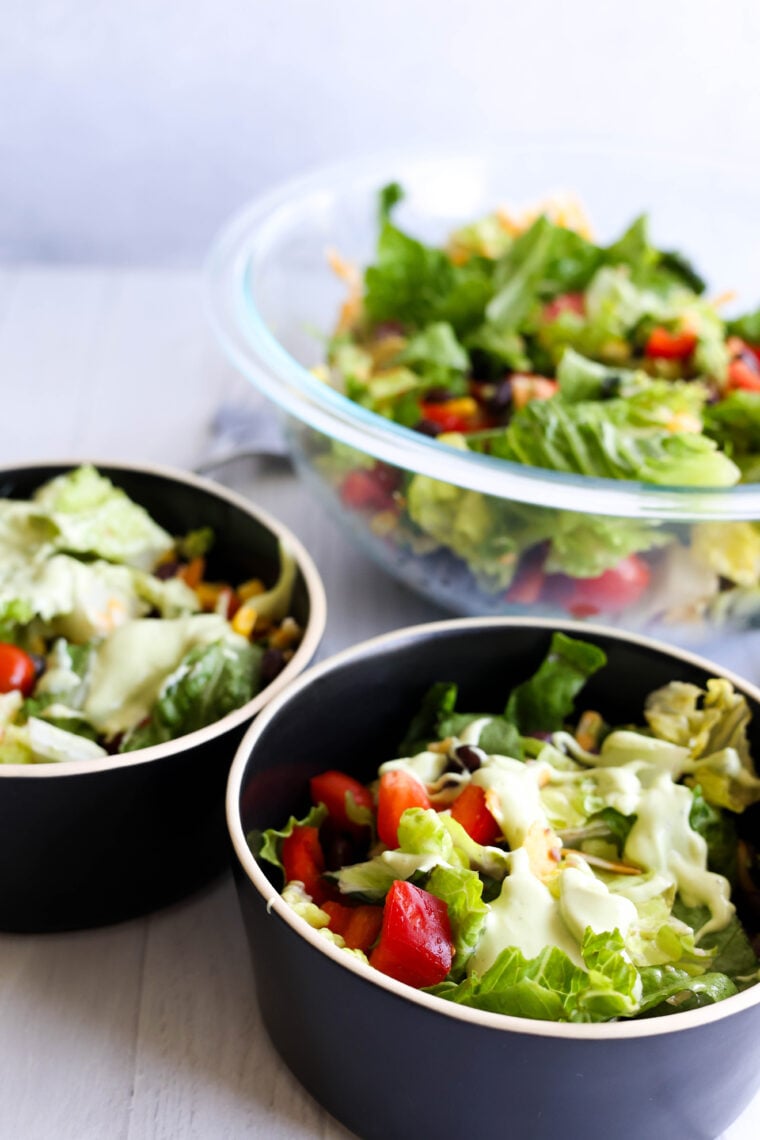 Three fresh salads with lettuce, tomatoes, and creamy dressing—two in small black bowls up front and a big glass bowl in back, all laid out on a light surface.