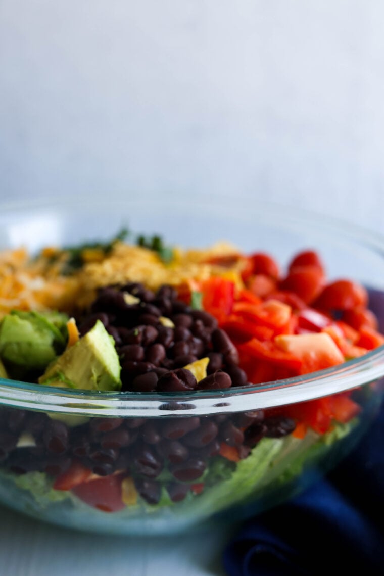 A close-up shot of a glass bowl packed with a vibrant salad—think black beans, chunks of avocado, diced and cherry tomatoes, plus shredded cheese on top.