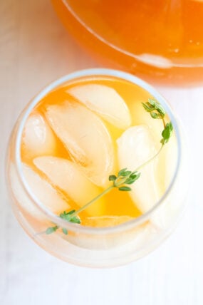 A close-up shot of a glass filled with an icy orange drink, topped with fresh thyme. You can see some ice cubes and part of the pitcher in the background.