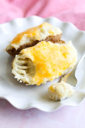 A fluffy baked potato split open, loaded with gooey melted cheddar, with a bite already taken and some cheesy goodness on a fork—all on a plain white plate.