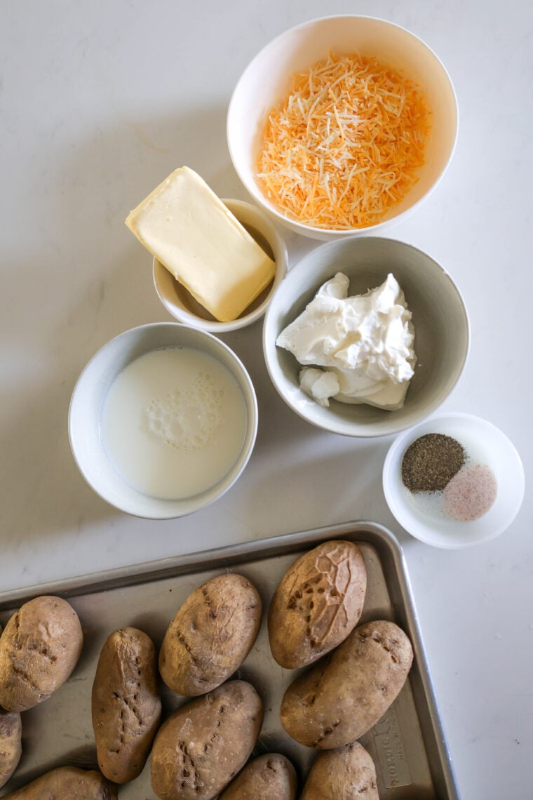 There’s a tray of baked potatoes on the counter, with bowls of shredded cheese, butter, milk, sour cream, and some salt and pepper nearby.