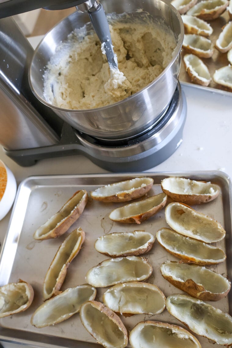 A metal mixing bowl filled with mashed potatoes sits next to trays of empty potato skins, all set and ready to be stuffed, right on the kitchen counter.