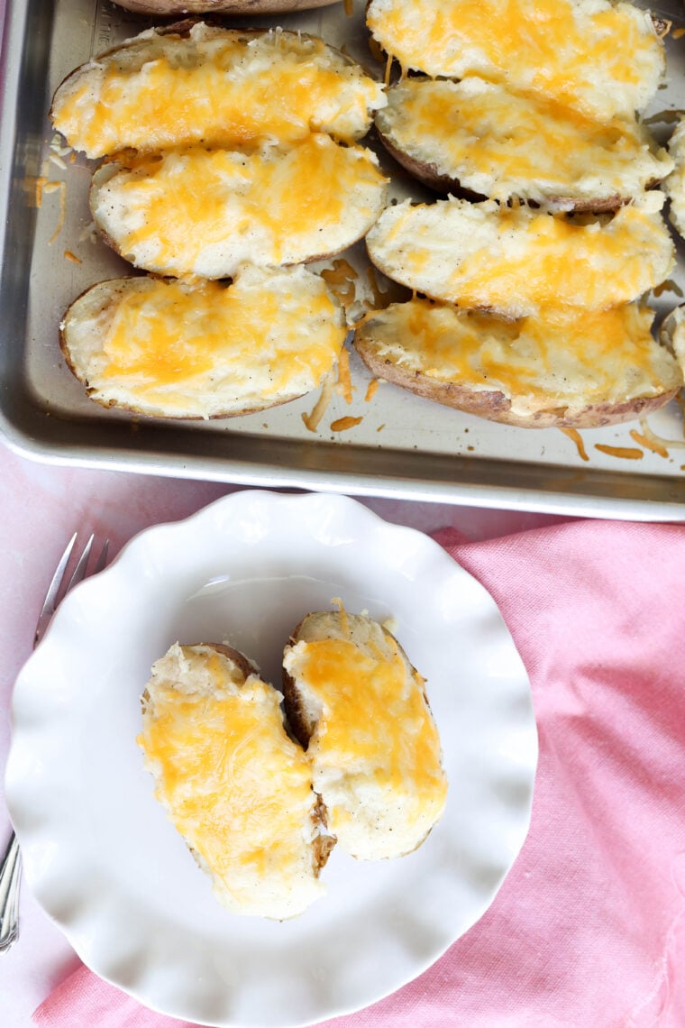 Cheesy twice-baked potatoes are lined up on a baking sheet, while two sit on a white scalloped plate over a pink cloth. Forks are right next to the plate, ready to dig in.