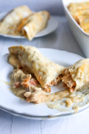 A close-up shot of cheesy chicken enchiladas on a white plate, smothered in creamy sauce. One’s cut open so you can see all that tasty filling, and there are more enchiladas hanging out in the background on another plate and in a baking dish.