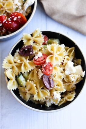 A bowl of Mediterranean pasta salad—packed with bow-tie pasta, cherry tomatoes, cucumber, feta, and Kalamata olives—sits on a white table. You can see another bowl peeking in from the corner.