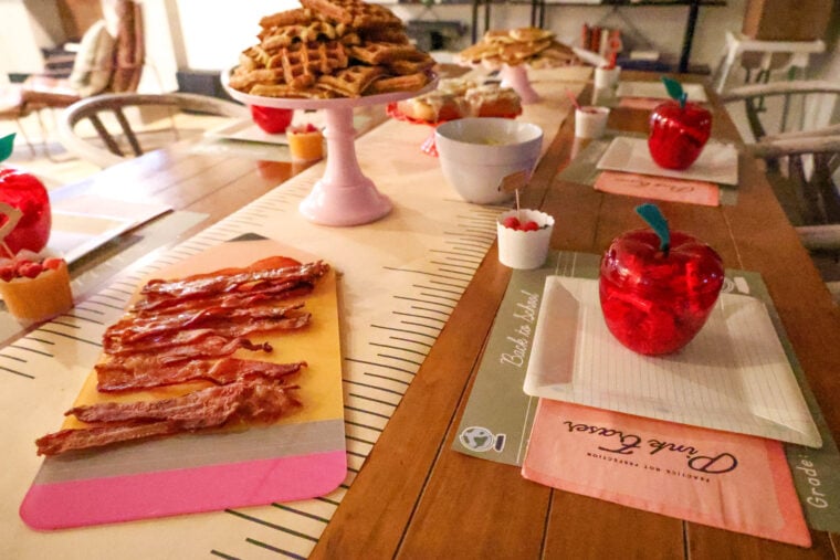 A wooden table’s all set for a cozy back-to-school breakfast—think bacon strips, waffles stacked on a cake stand, cups of berries, and cute apple-shaped containers on notebook paper placemats. The whole vibe is super warm and welcoming.