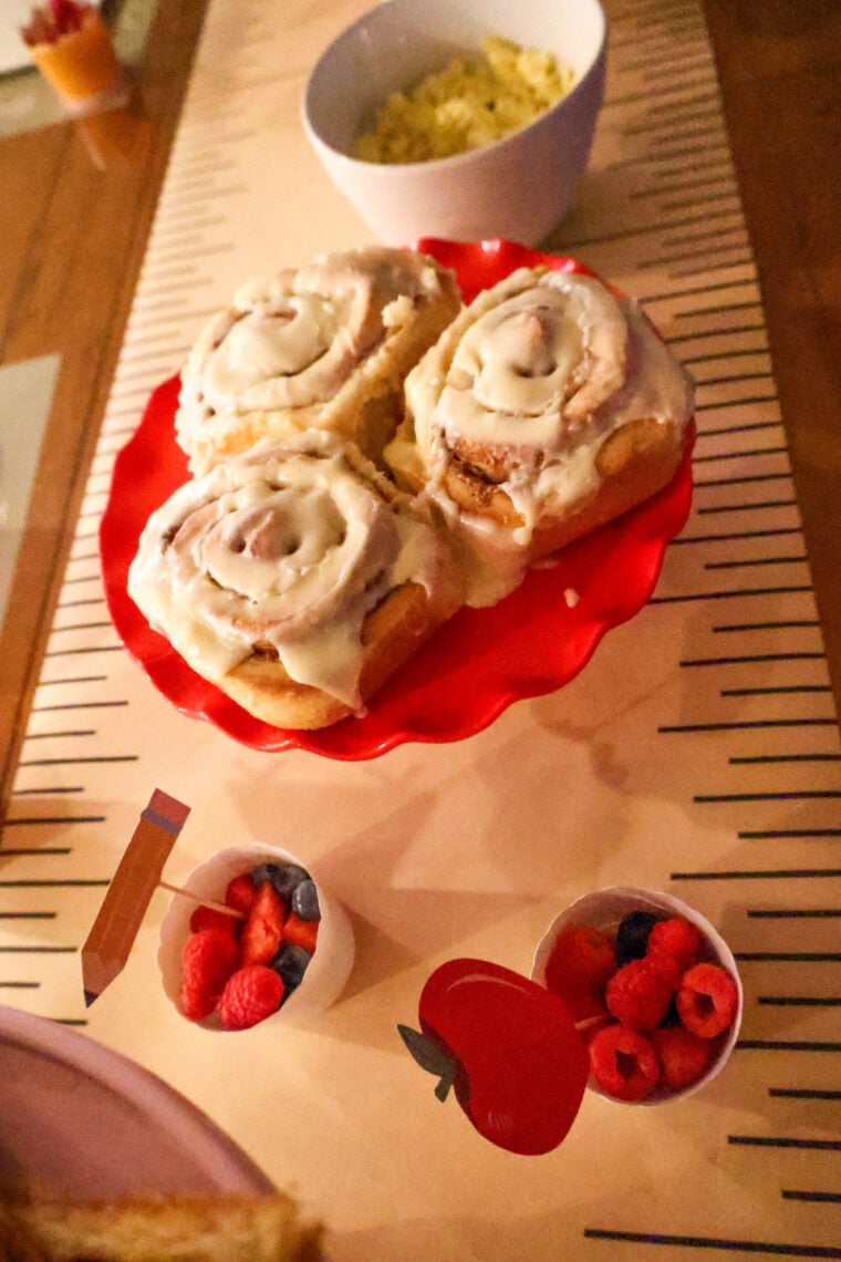 Three frosted cinnamon rolls on a red cake stand, scrambled eggs in the back, and two cups of fresh berries up front—makes for an easy and tasty back-to-school breakfast!.