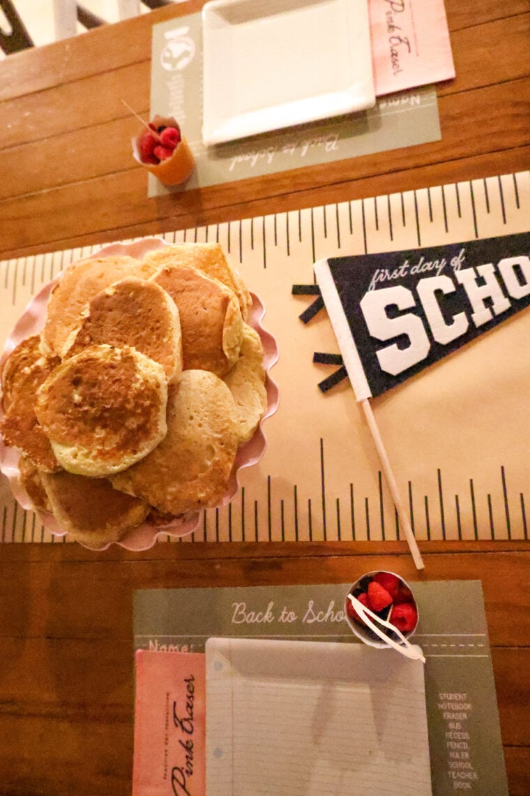 A stack of pancakes is ready on the table, surrounded by school-themed placemats, a “first day of school” flag, some berries in little cups, and blank notepaper with pencils—making breakfast totally feel like a back-to-school party.