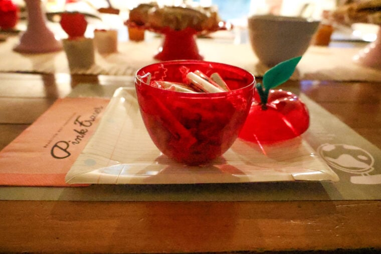 Theres a red, apple-shaped bowl full of wrapped candies on a white tray, sitting on a wooden table and all set for a back-to-school breakfast. In the background, you can spot a pink napkin and some other blurred stuff.