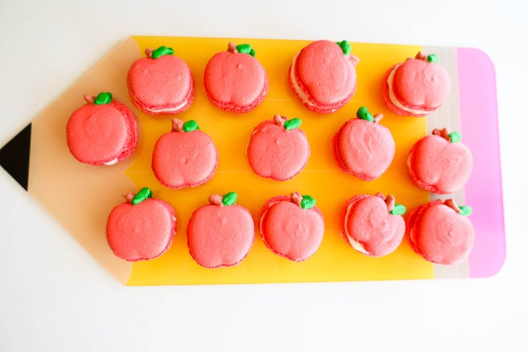 Pink macarons made to look like apples—with cute green leaves and brown stems—are lined up on a big pencil-shaped board in yellow and pink. It’s the perfect, festive setup for a back-to-school breakfast!.