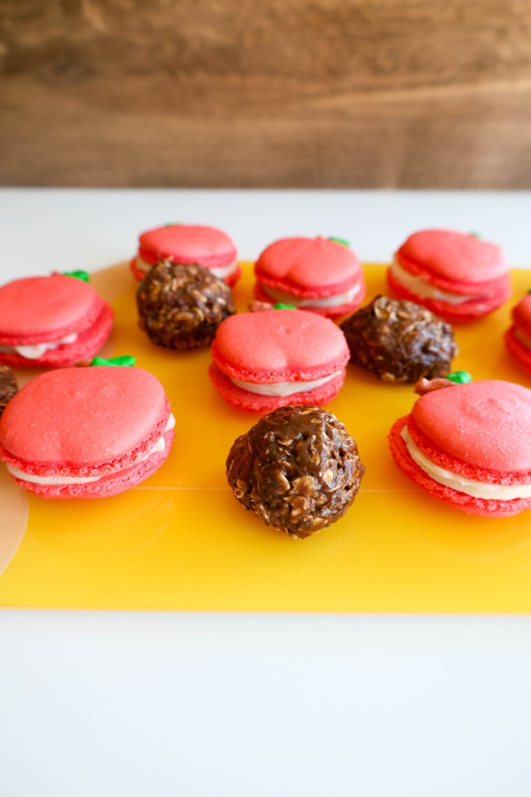 A bunch of pink macarons with creamy filling and green sprinkles are set out on a yellow surface, right next to some round, chocolatey treats. They’re perfect for kicking off your back-to-school morning!.