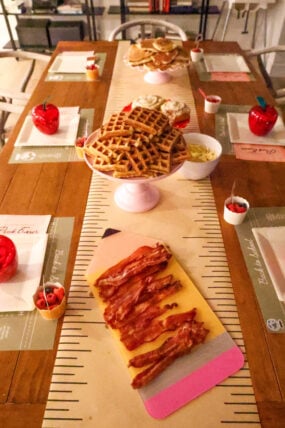 A wooden tables all set for a back-to-school breakfast with waffles, bacon, cupcakes, and bowls of toppings. Each seat has a placemat and there are cute red apple decorations. Stacks of waffles and cupcakes on cake stands make the whole thing feel super festive!.