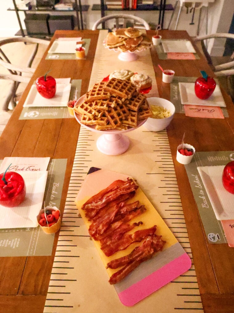 A wooden tables all set for a back-to-school breakfast with waffles, bacon, cupcakes, and bowls of toppings. Each seat has a placemat and there are cute red apple decorations. Stacks of waffles and cupcakes on cake stands make the whole thing feel super festive!.