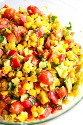 A close-up shot of a colorful charred corn salad packed with cherry tomatoes, avocado, red bell pepper, and cilantro, all tossed together with a light dressing in a glass bowl.