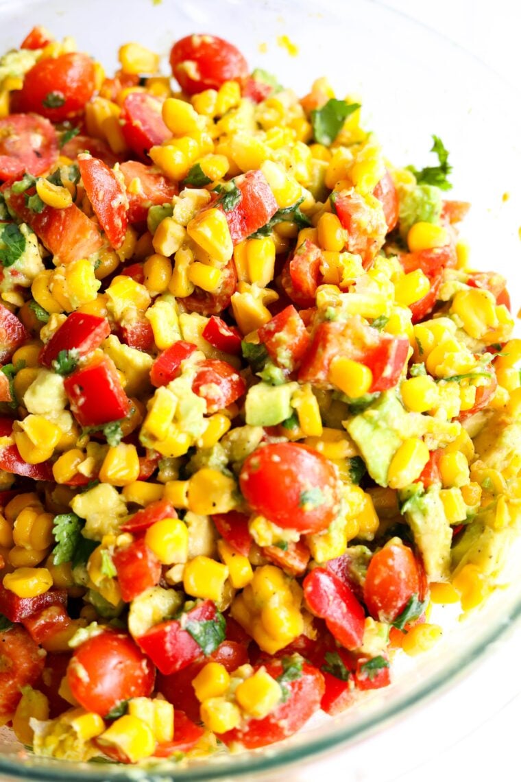 A close-up shot of a colorful charred corn salad packed with cherry tomatoes, avocado, red bell pepper, and cilantro, all tossed together with a light dressing in a glass bowl.
