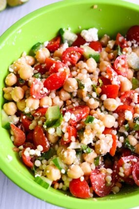 A green bowl packed with colorful couscous salad—chickpeas, cherry tomatoes, green onions, feta, and fresh herbs all mixed in.
