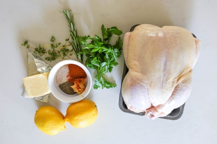 A whole spatchcock chicken is laid out on a tray with fresh herbs, a couple of lemons, some butter, and a little bowl of spices and minced garlic—all ready to go on a clean white countertop.