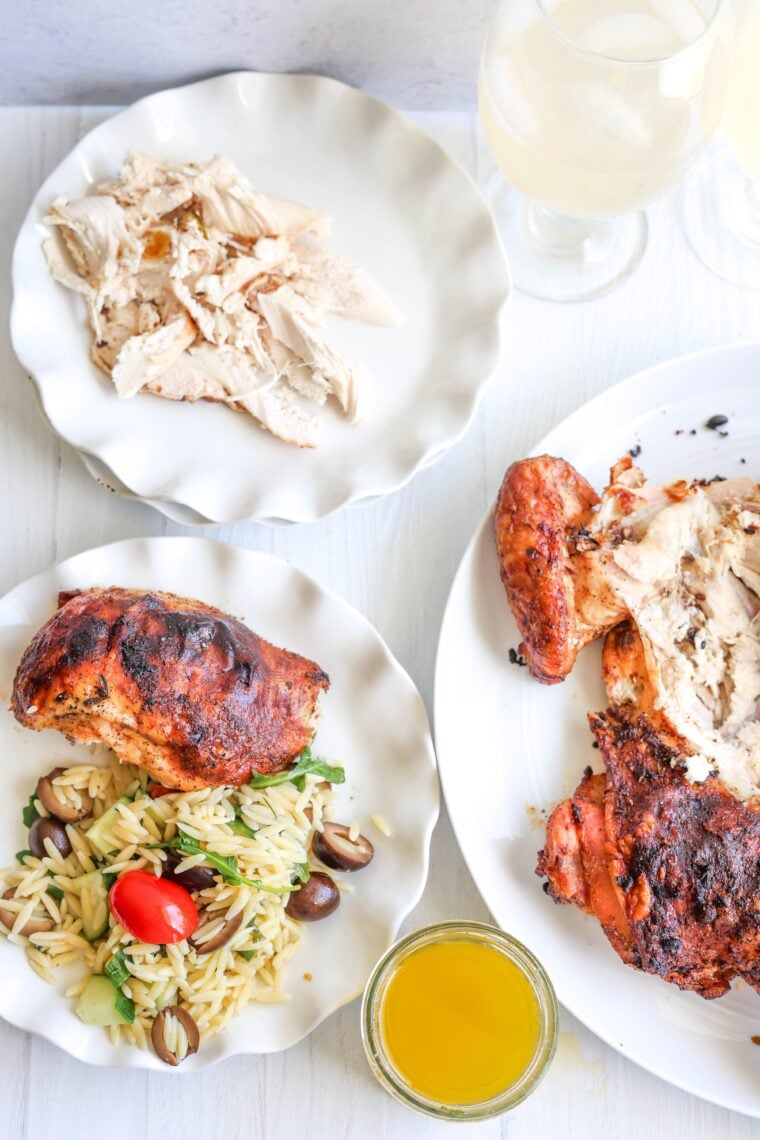 There are three white plates on the table—one’s got shredded chicken, another has a spatchcock chicken with orzo pasta salad and veggies, and the last one has a partly carved roast chicken. Plus, there’s a glass of something yellow and two wine glasses nearby.