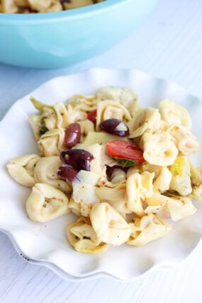 A white scalloped plate loaded with tortellini pasta salad, topped with artichoke, olives, and cherry tomatoes, sitting on a light surface.