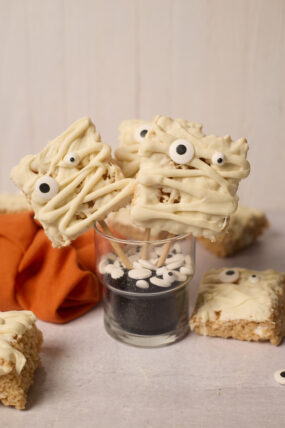 Mummy Rice Krispies with white chocolate and candy eyes on sticks sit in a sprinkle-filled glass. Extras chill on an orange napkin.