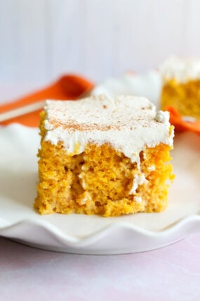 A square piece of pumpkin tres leches cake with creamy frosting and a sprinkle of cinnamon sits on a white scalloped plate.