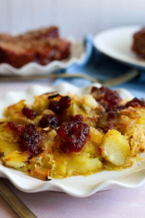 Close-up of crispy smashed potatoes with gooey brie and cranberry sauce, plus some blurred meatloaf slices in the back.