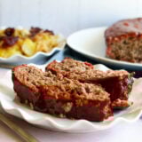 Two juicy slices of smoked meatloaf with a sweet glaze sit on a plate with roasted potatoes, plus more meatloaf behind them.