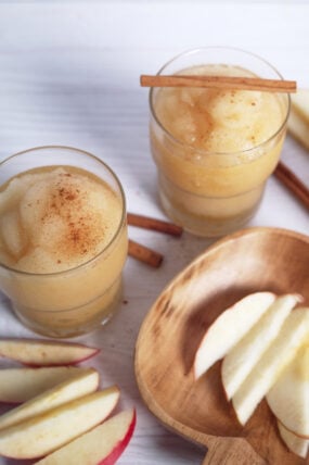 Two glasses of apple cider slush with cinnamon and cinnamon sticks next to a wooden plate of apple slices on a white table.