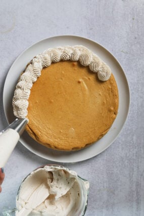 Someone is piping white cinnamon whipped cream onto the edge of a golden cake on a plate, frosting bowl close by.