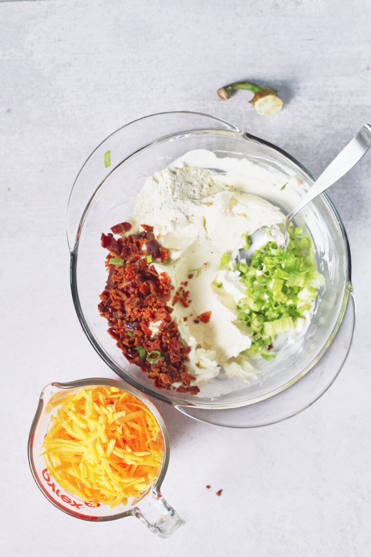 Cream cheese, flour, green onions, and bacon fill a bowl with a spoon. Cheddar waits in a cup nearby for pumpkin cheeseball fun.