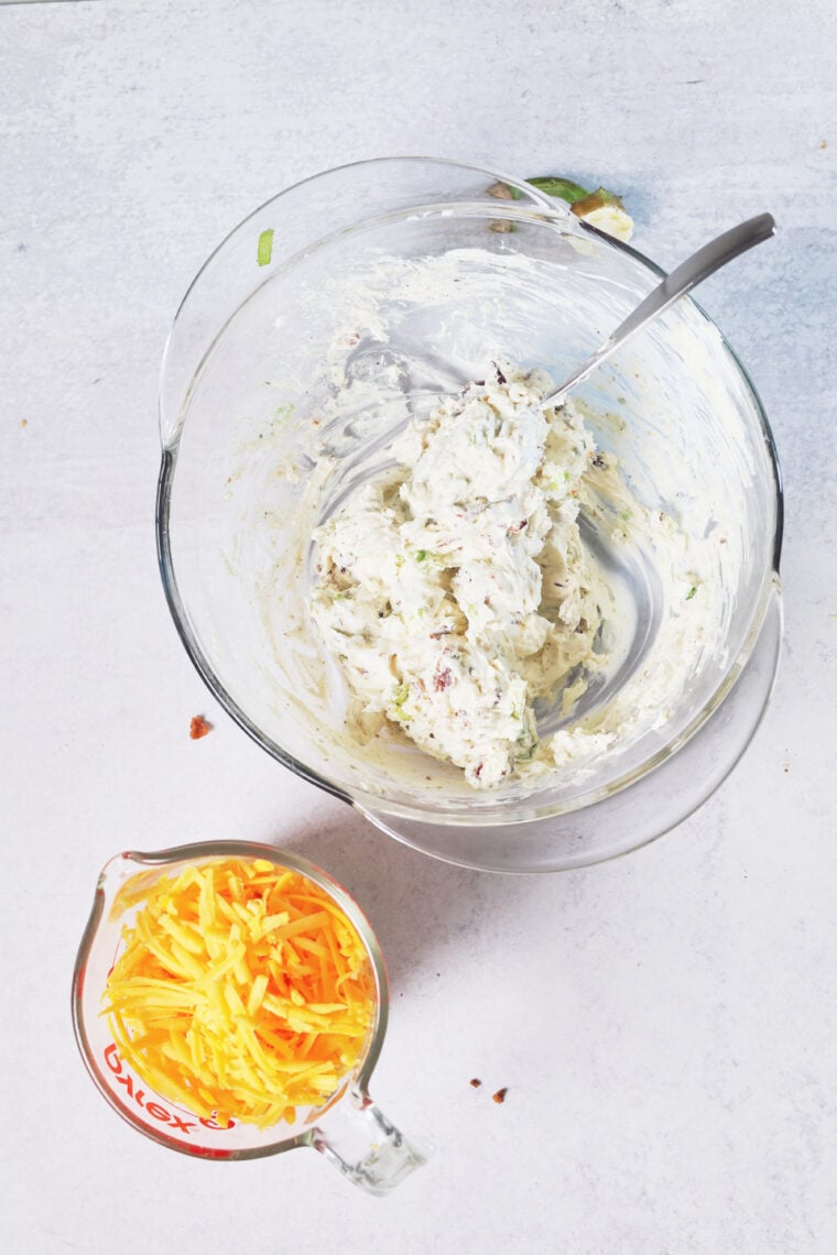 There’s a glass bowl with creamy pumpkin cheeseball mix and a spoon, chilling next to a measuring cup packed with shredded cheese.