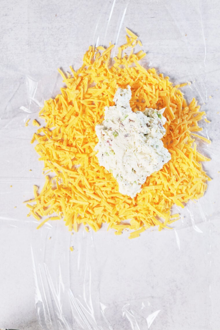 Shredded cheddar and herbed cream cheese on plastic wrap, prepped for a tasty pumpkin cheeseball, on a pale countertop.