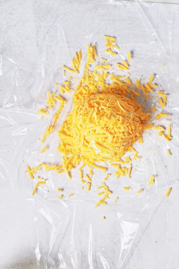 A pile of shredded cheddar cheese sits on clear plastic wrap, all set to be turned into a pumpkin cheeseball.