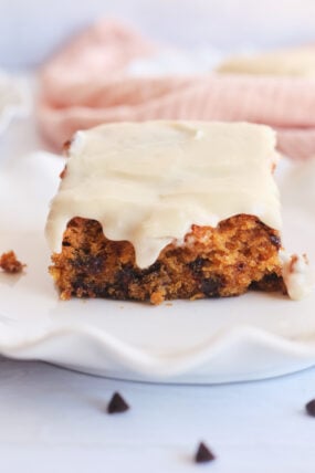 A moist square of pumpkin chocolate chip cake with creamy white frosting sits on a scalloped white plate, up close.
