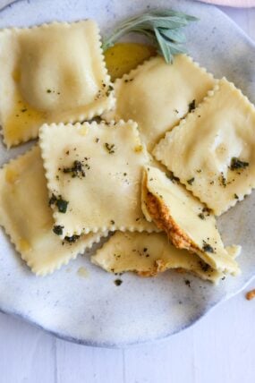 Square pumpkin ravioli on a plate with browned butter, herbs, and fresh sage. One’s cut open to show the creamy orange inside.