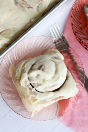 A cinnamon roll with creamy icing sits on a pink glass plate, right next to a pan full of more frosted rolls. There’s also a pink napkin and fork ready to go.
