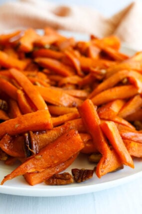 Roasted carrot sticks, sweet potato wedges, and pecans on a plate, all golden and caramelized, with a beige cloth behind.